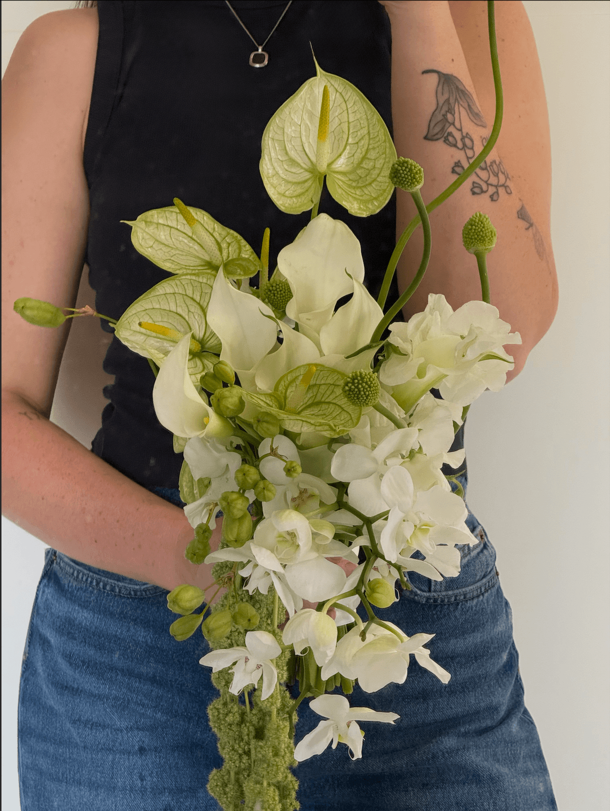 Person holding a bouquet of white flowers with green leaves, wearing a black sleeveless top and blue jeans.