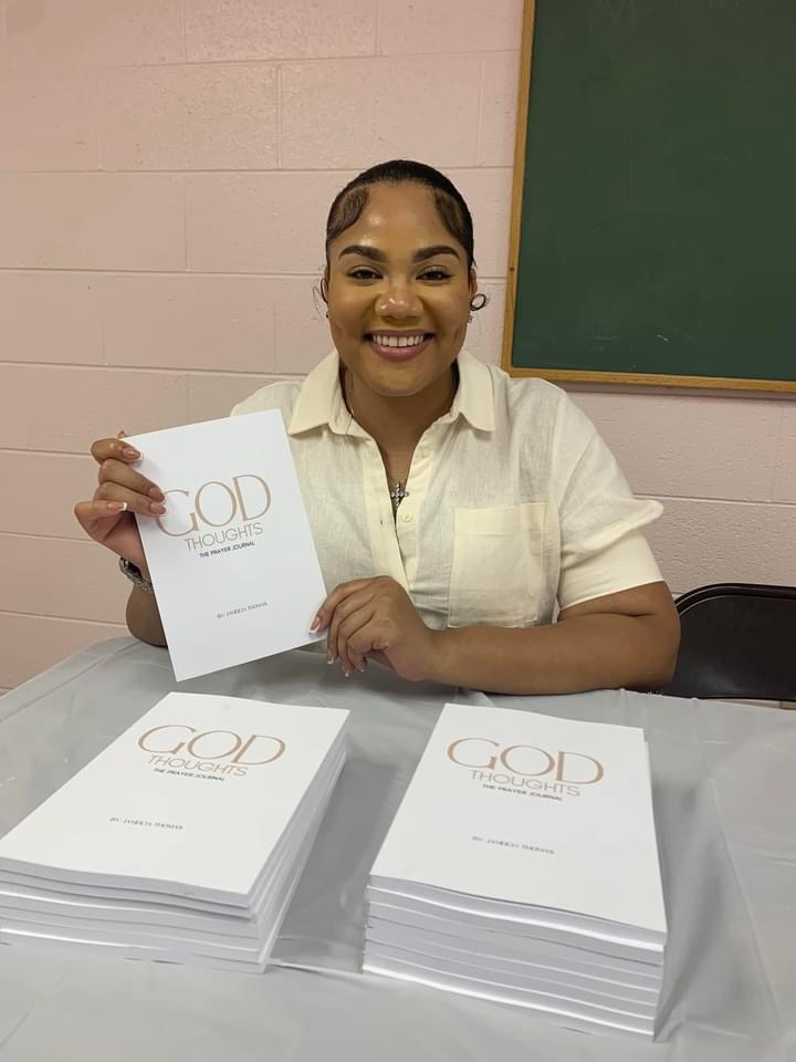 Smiling woman sitting at table holding a booklet, with stacks of similar booklets in front, classroom setting with green chalkboard behind.