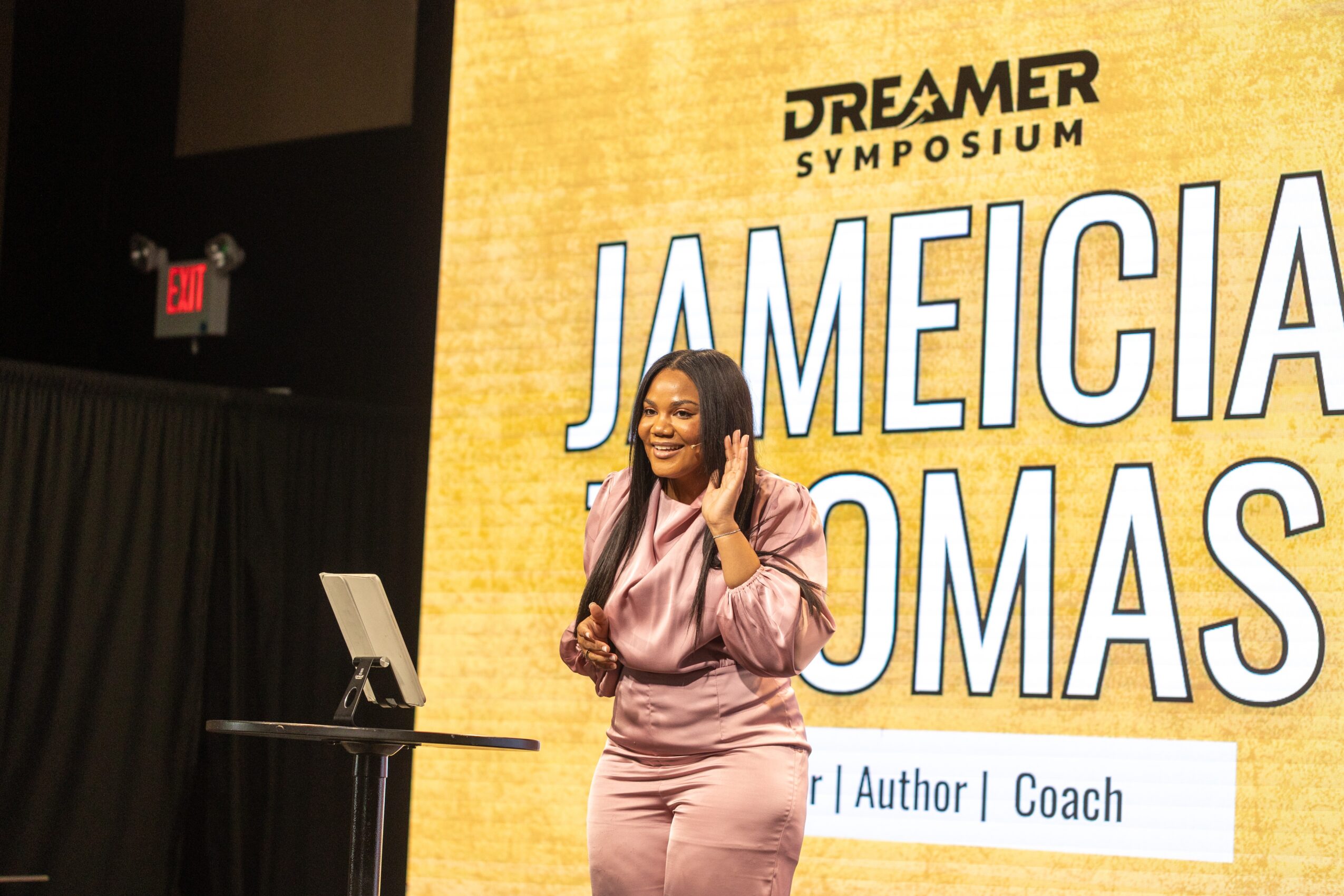 Woman speaking on stage at Dreamer Symposium with large screen behind her displaying her name and title.