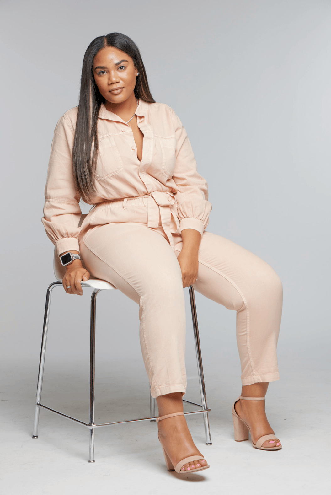 Woman sitting on a stool, wearing a beige outfit and high heels, against a plain background.