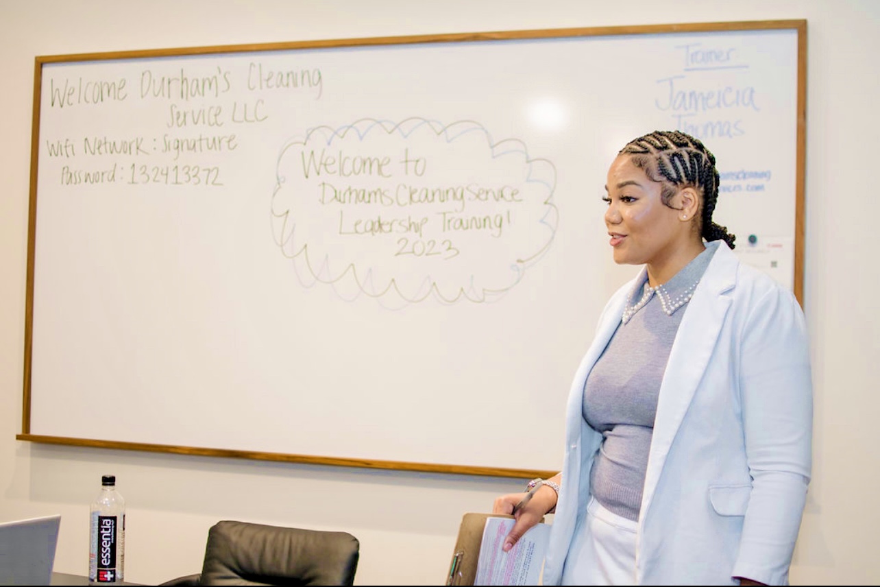 Woman standing in front of a whiteboard with writing, wearing a light blue blazer and gray top, smiling.