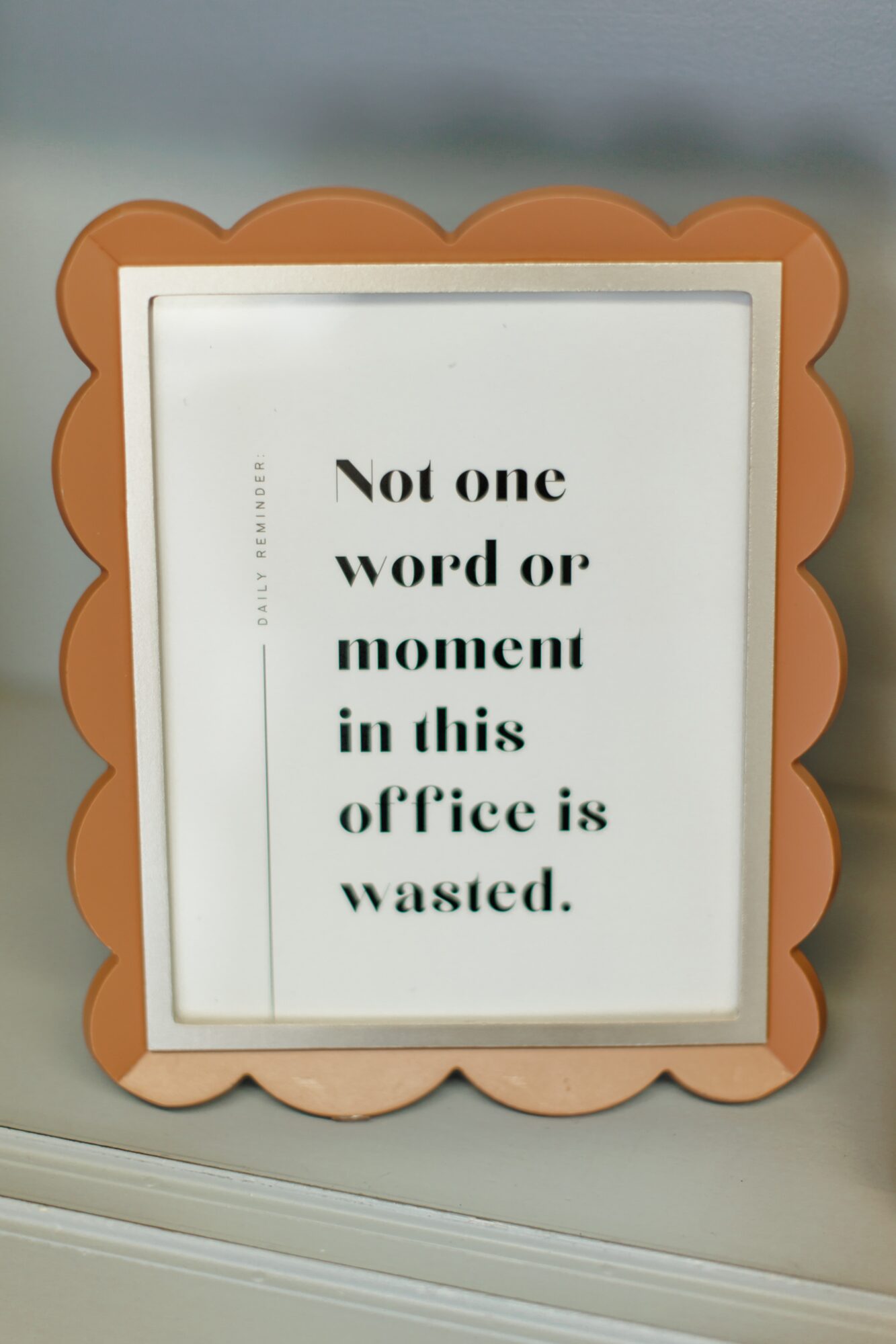 Sign with text: 'Not one word or moment in this office is wasted.' framed with a scalloped orange border.
