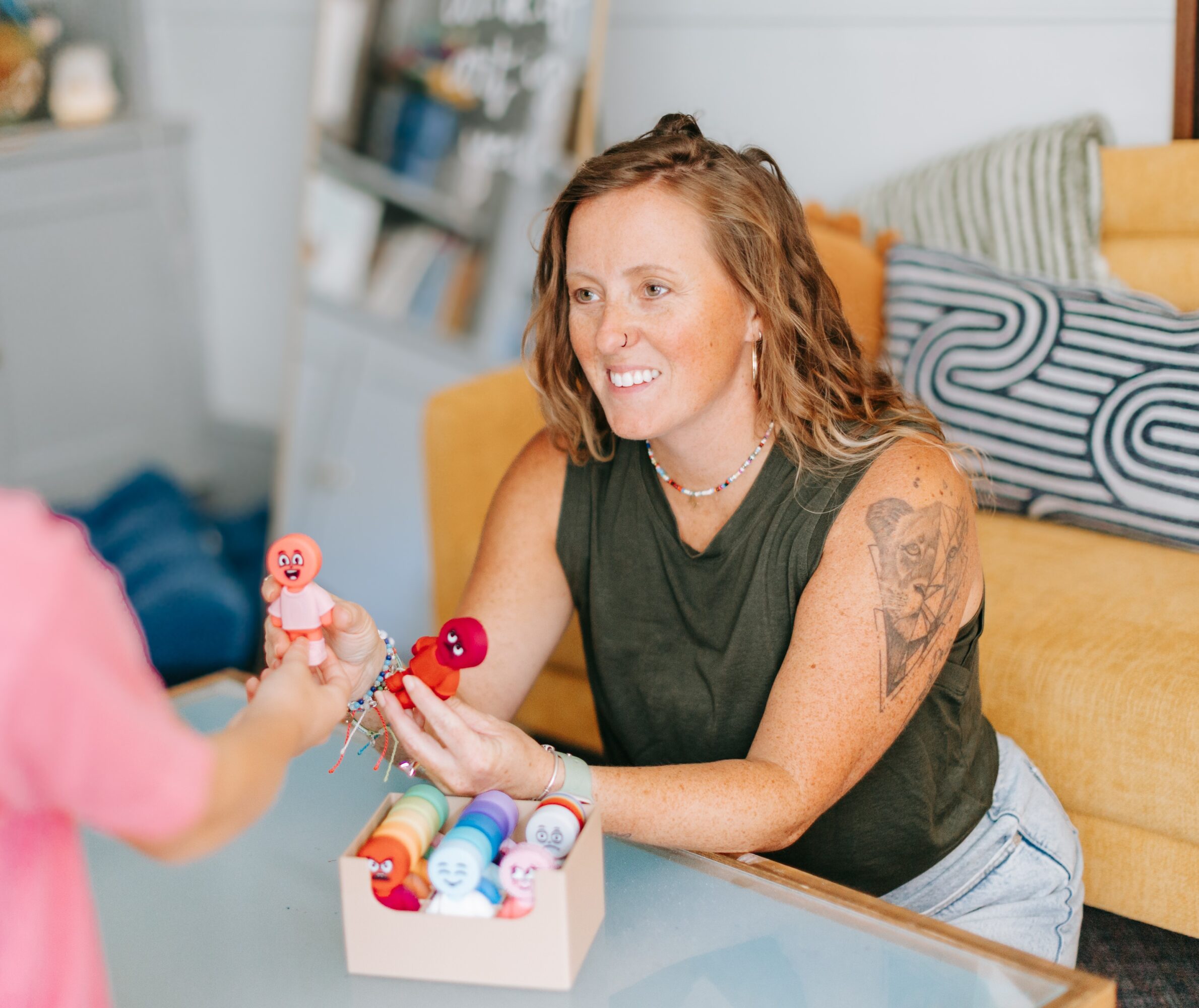 Woman with curly hair and tattoos smiling at a child, playing with dolls on a table in a living room.