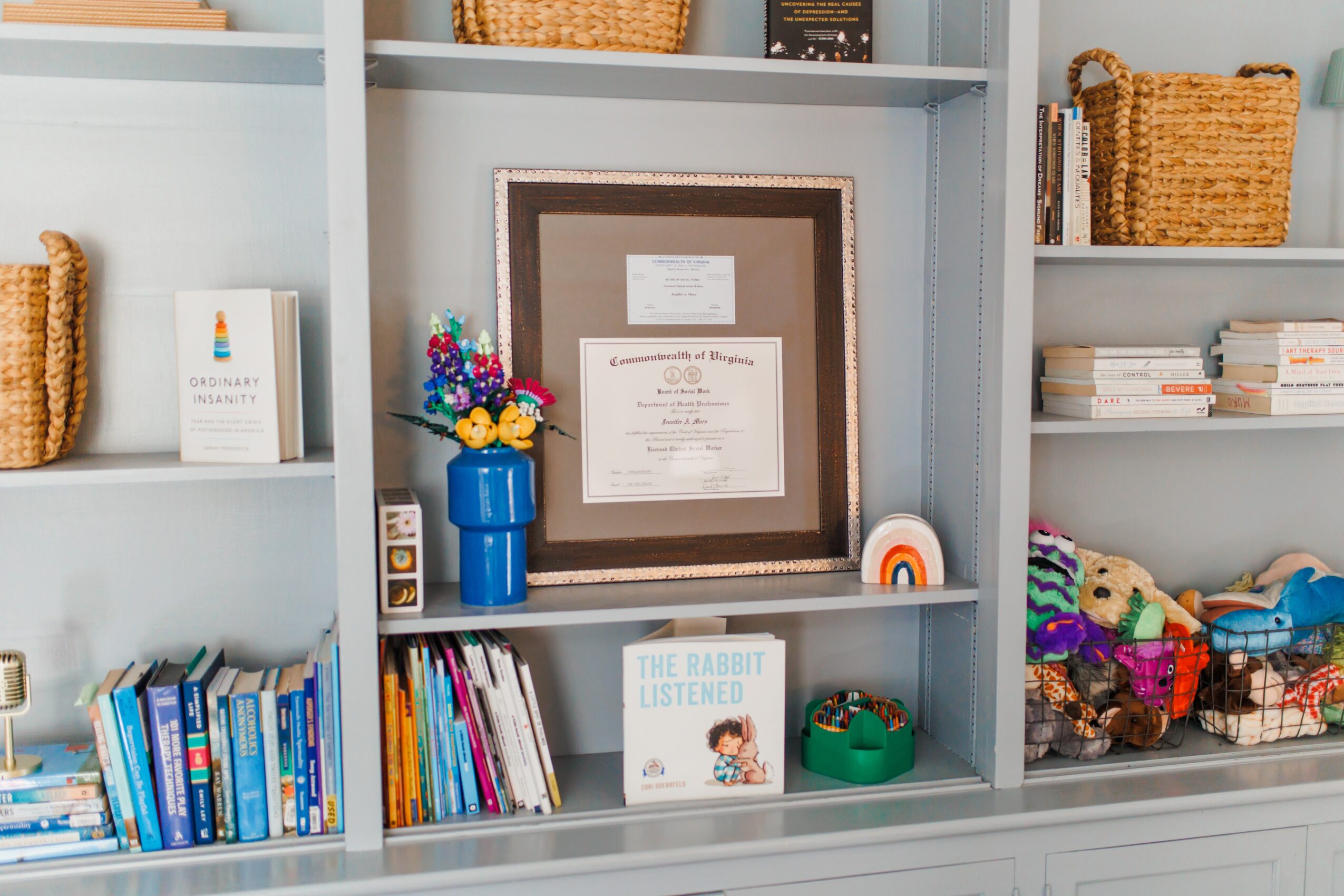 Bookshelf with framed certificate, colorful flowers in a blue vase, and various books and baskets.