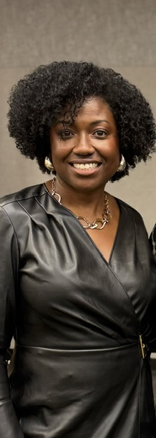 Woman with curly hair smiling, wearing a black satin dress and a necklace, against a neutral background.