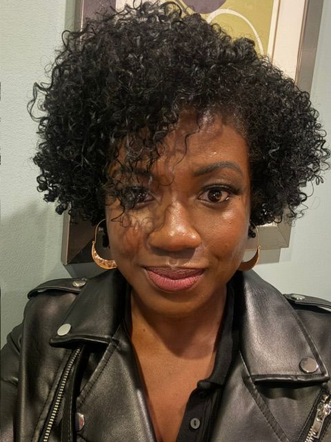 Woman with curly hair wearing a black leather jacket, smiling, indoors with a framed picture in the background.