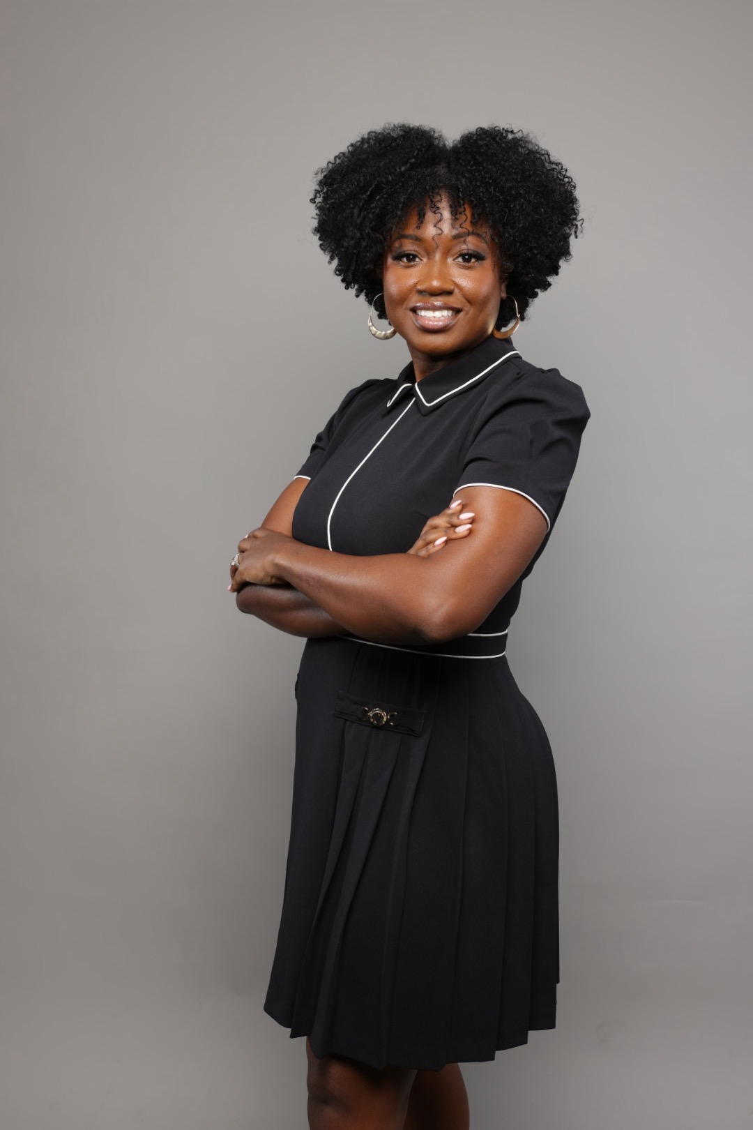 Young woman with curly hair smiling, arms crossed, wearing a black dress with white piping, standing against a gray background.