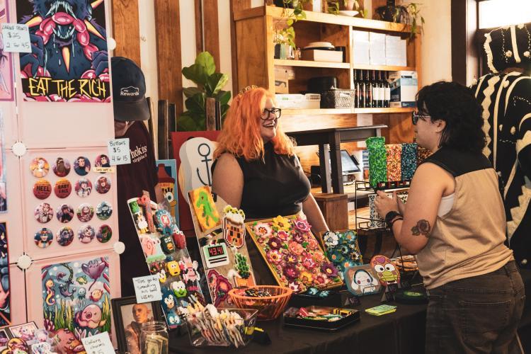 Two women with dark hair and glasses converse at a colorful craft booth inside a store, surrounded by art and accessories.