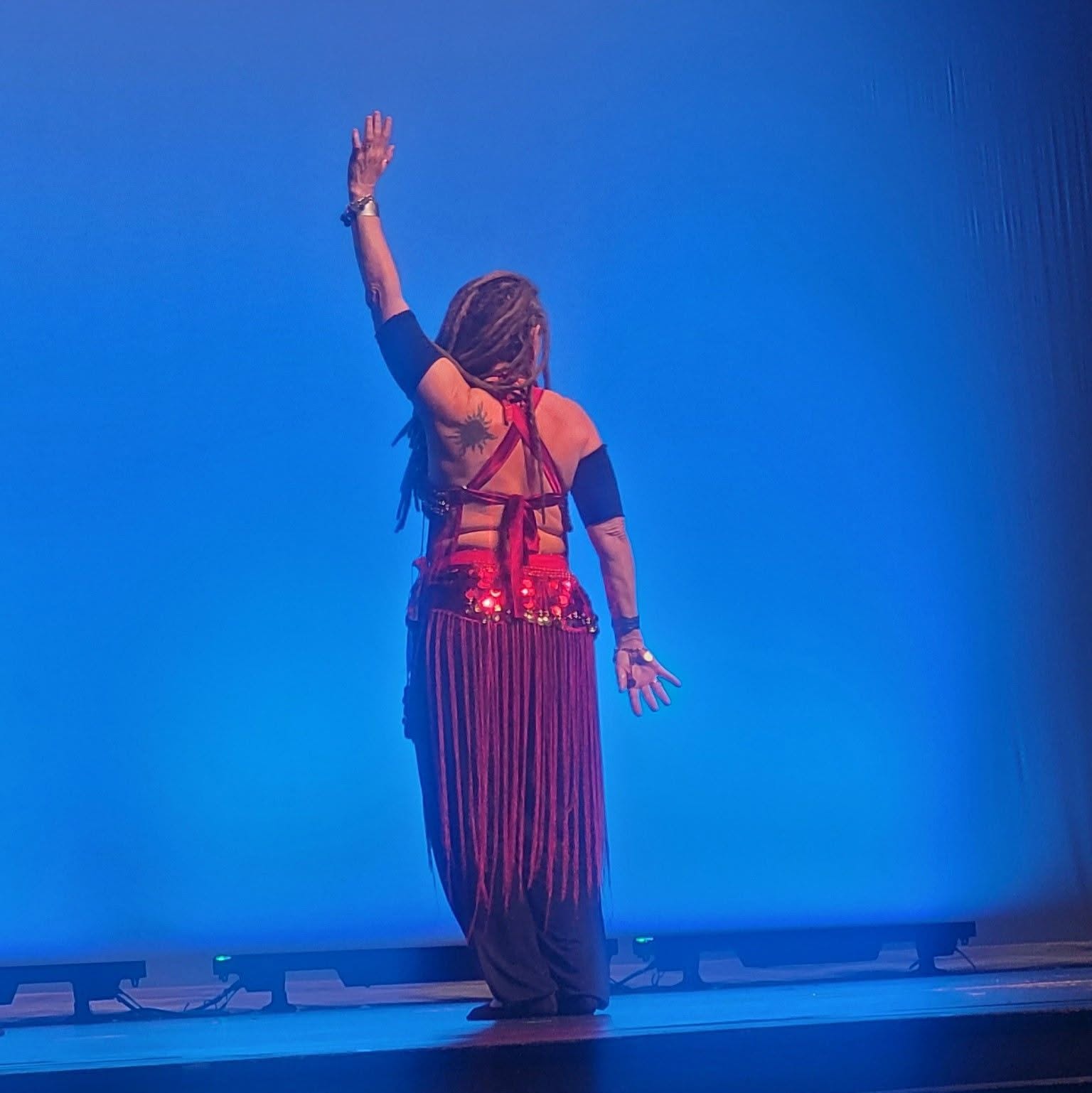 Person with long hair in a colorful outfit, standing on stage with one arm raised, facing away from the camera.