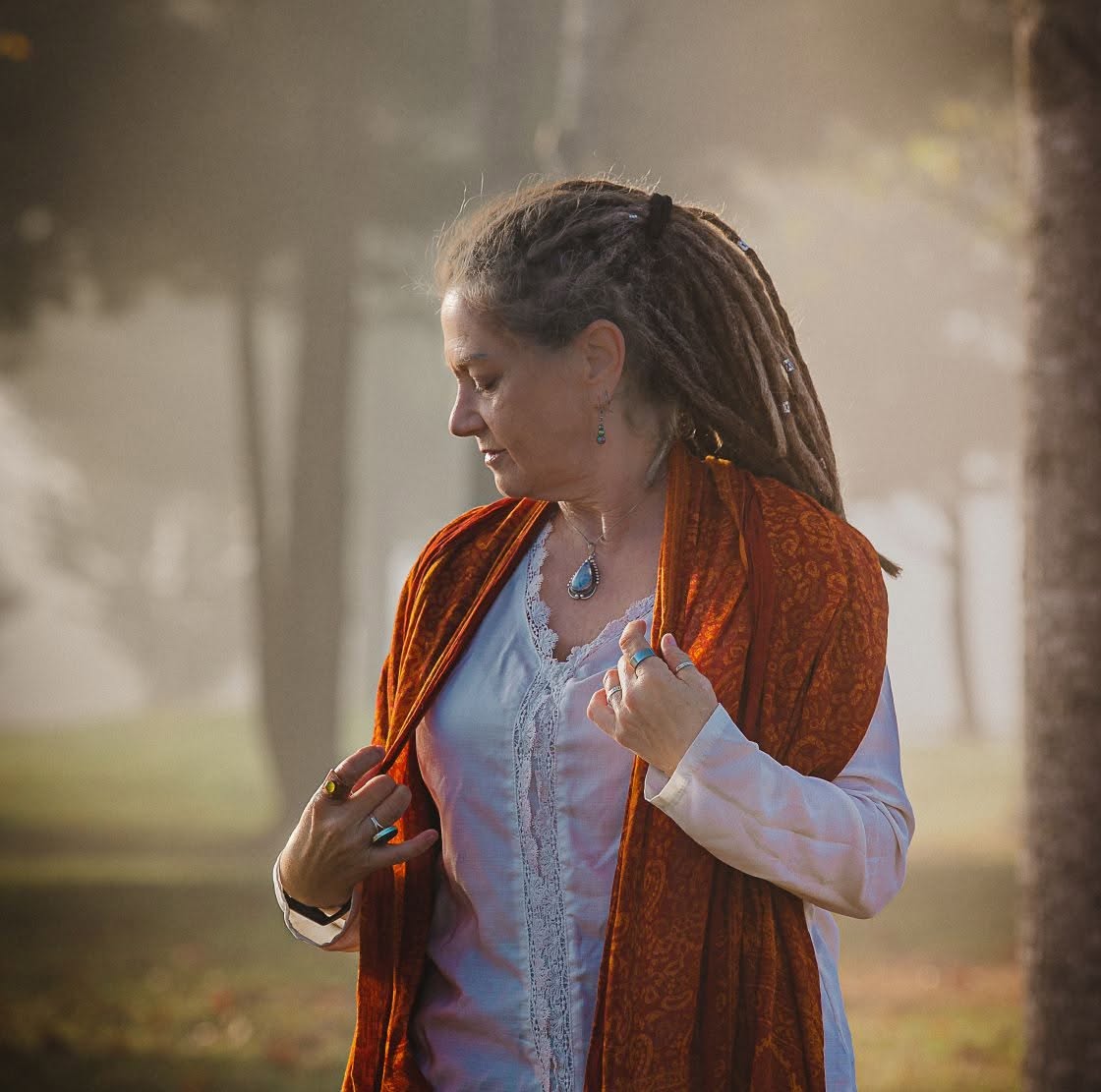 Woman with dreadlocks wearing a white blouse and orange shawl, standing outdoors in a park with trees and sunlight.