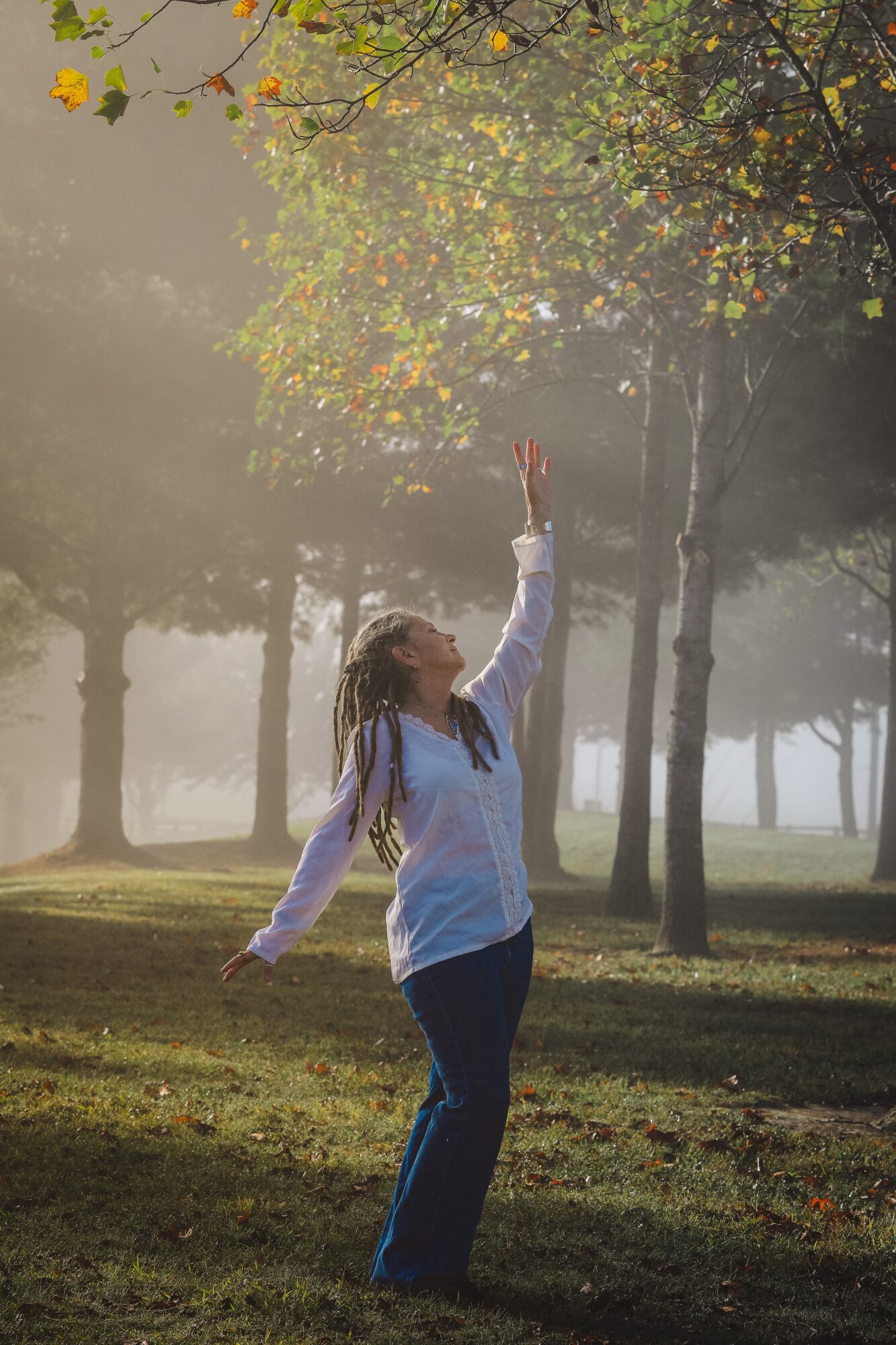 Child with long hair reaching up towards tree branches in a park during foggy morning.