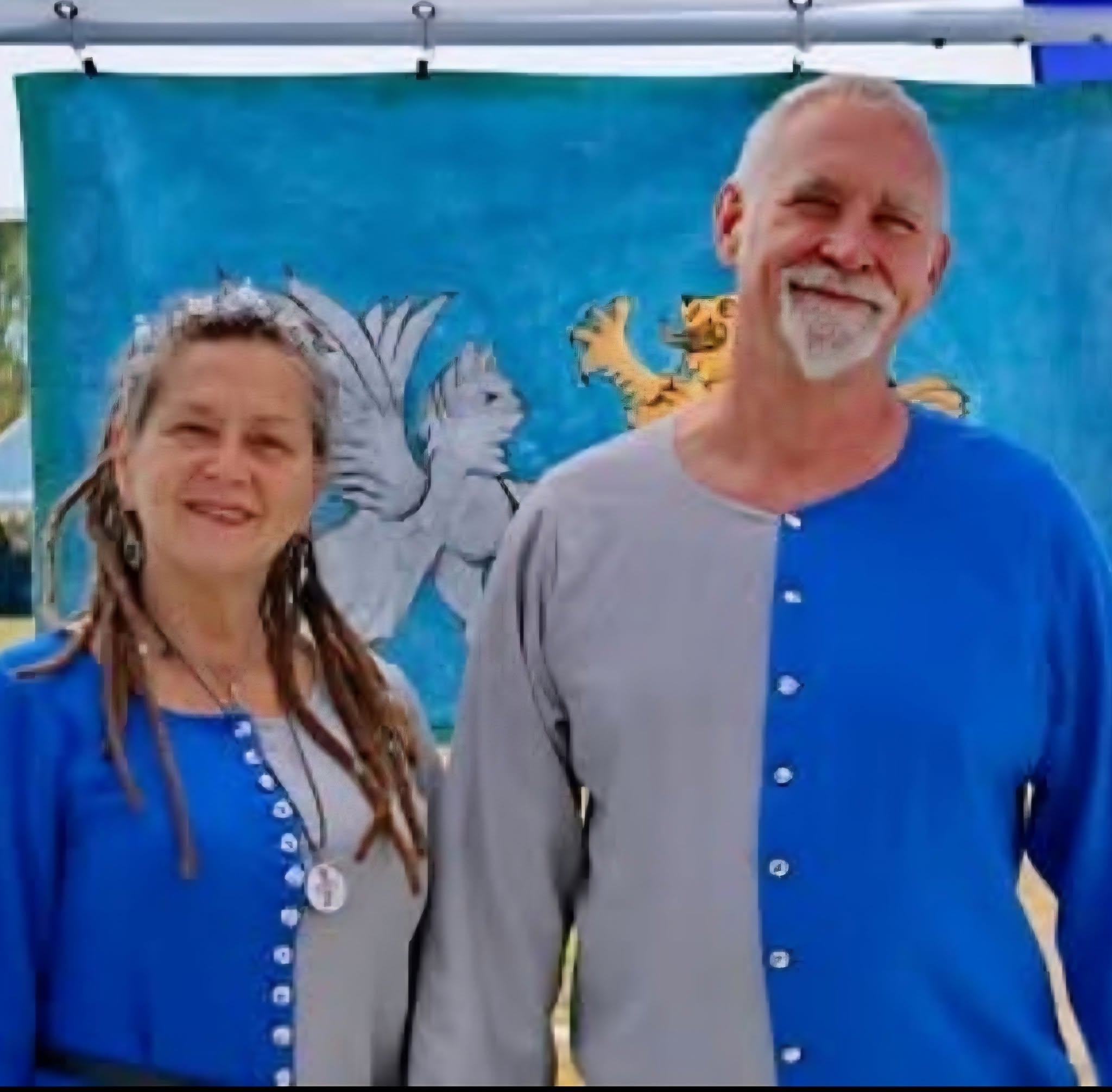 Two people, a woman and a man, stand outdoors in front of a colorful backdrop with a dragon and a tiger. They wear matching blue and gray clothing, smiling.