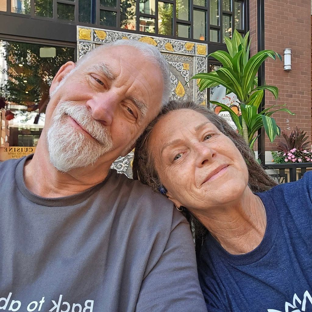 Two older adults smiling and leaning their heads together outdoors with plants and a building in the background.