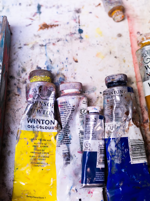 Several used paint tubes on a paint-splattered surface, with some tubes partially squeezed and paint residue visible.