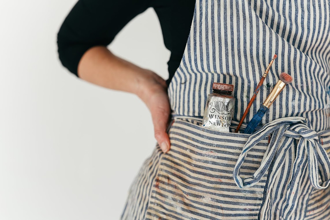 Person wearing a striped apron with art supplies in the pocket, hand on hip, black shirt visible, plain background.