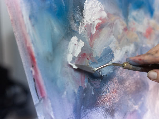 Close-up of a person using a palette knife to paint on a canvas with abstract colors and textures.