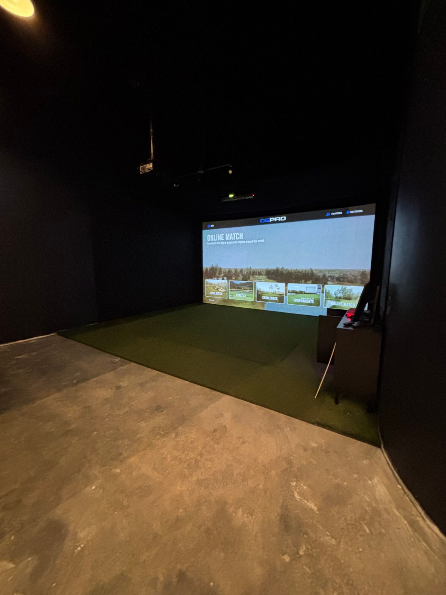 Indoor golf simulator room with a large screen displaying a golf course, a golf club, and a mat on the floor.