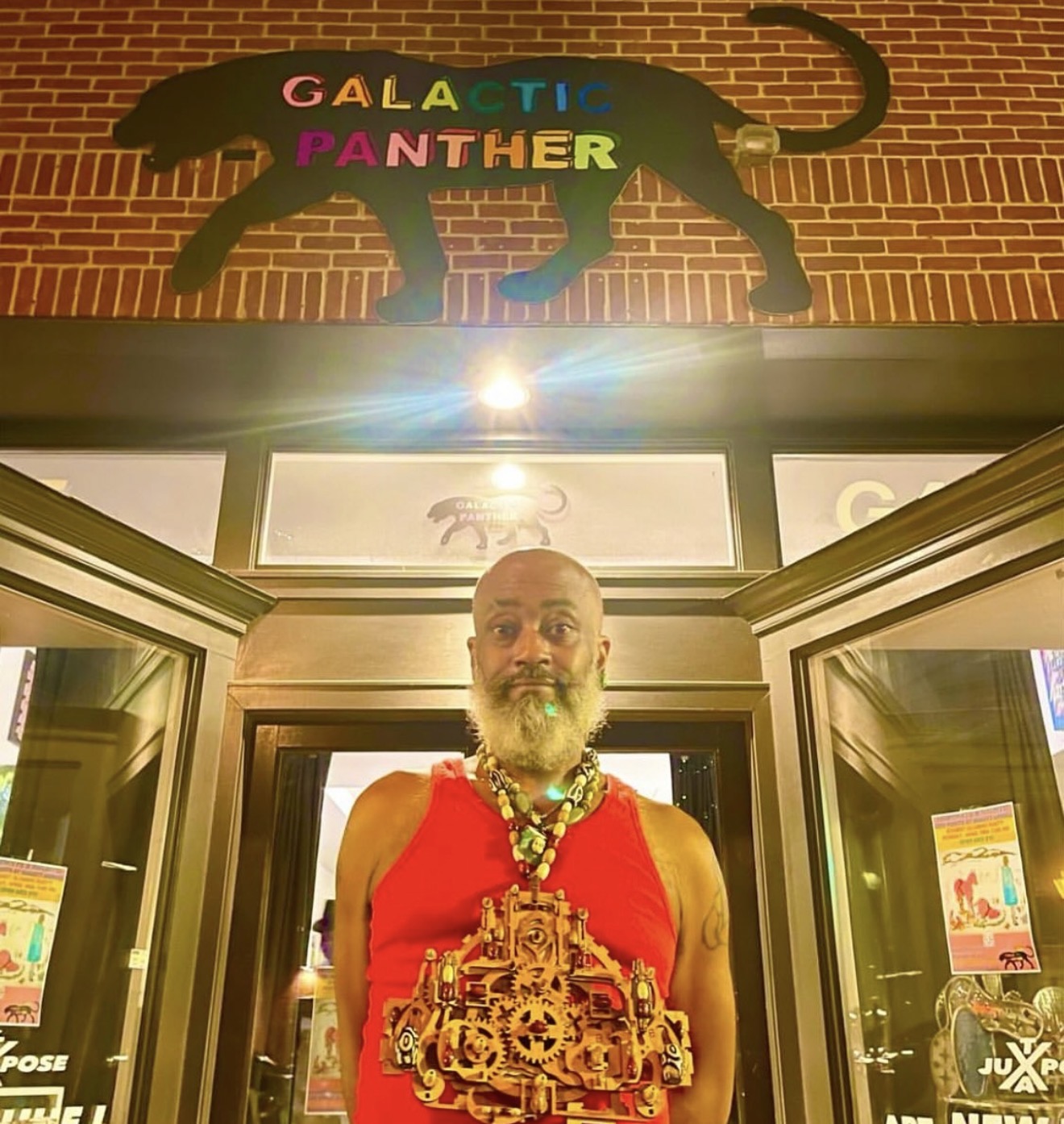 Man with a beard and necklace standing in front of a building entrance with a sign of a black panther and colorful text.
