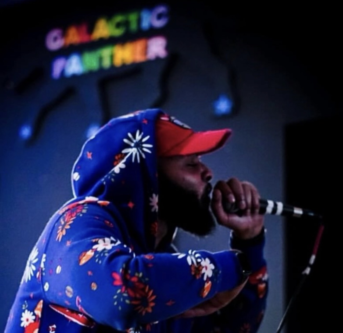 Person wearing a colorful hoodie and red cap, holding a microphone, with a sign reading 'GALACTIC PARTNER' in rainbow colors behind them.