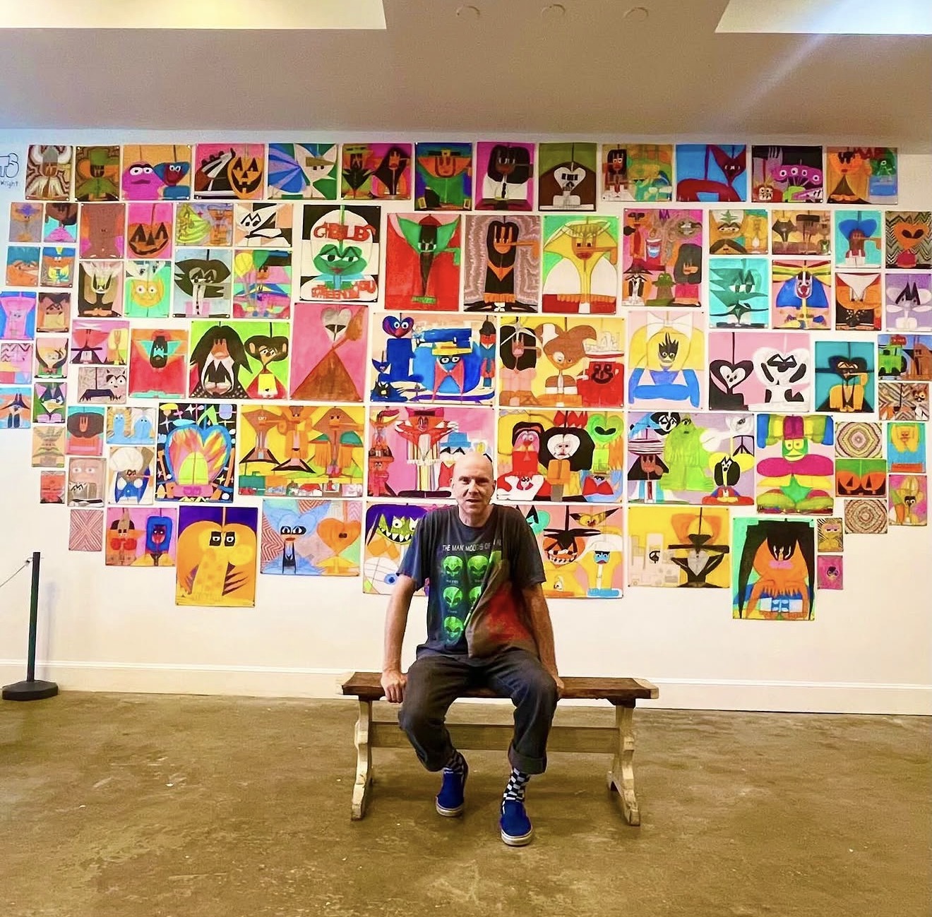 Person sitting on a bench in front of a colorful wall of artwork, with various vibrant paintings displayed behind.