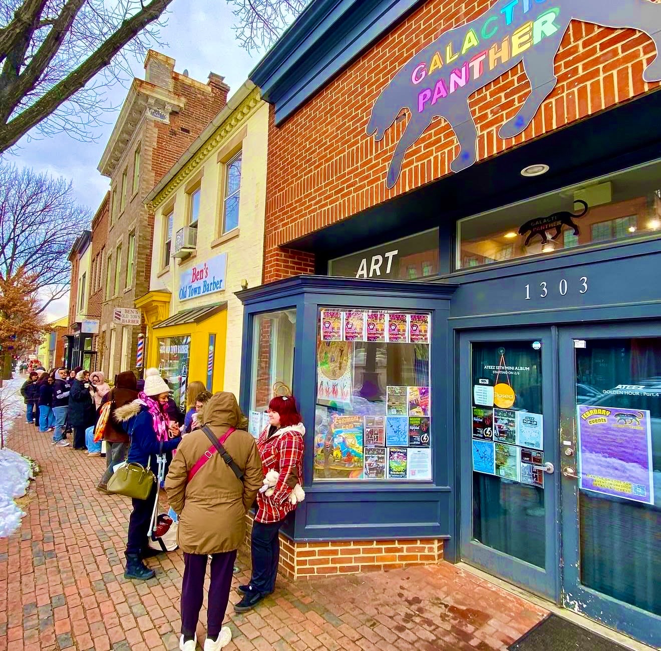 People line up outside a colorful art gallery on a brick sidewalk, with buildings and trees in the background.