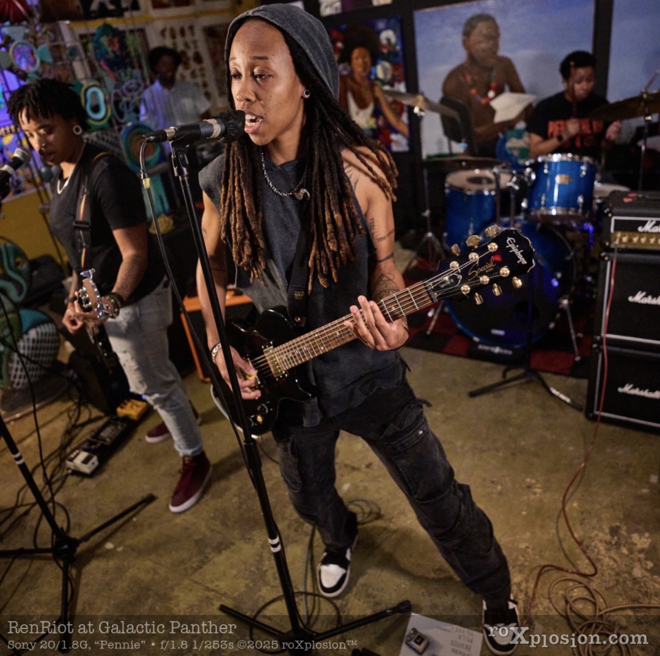 Group of musicians performing with guitars and drums in a colorful room, with a woman singing into a microphone.