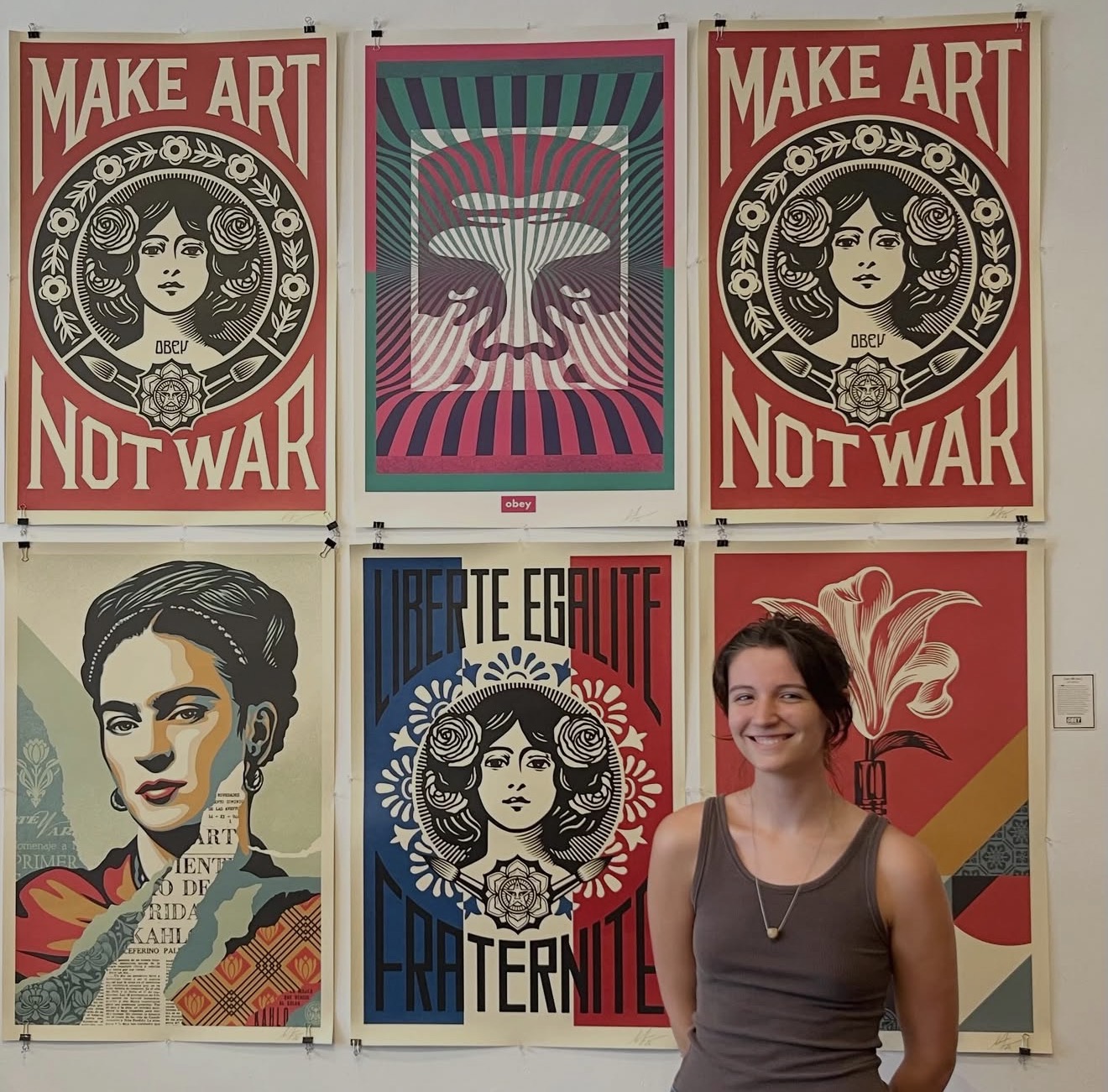 Young woman with short hair and a necklace standing in front of colorful artwork posters, smiling.