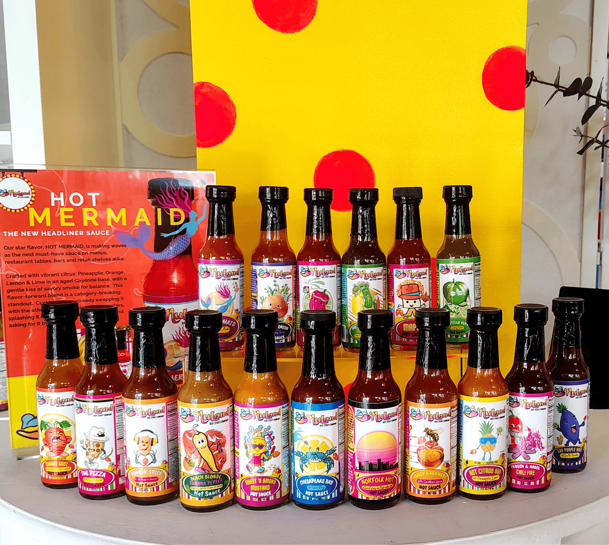 Collection of hot sauce bottles with colorful labels arranged on a table, with a yellow background featuring red dots.