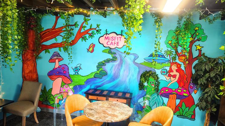 Colorful mural of fairytale characters, trees, and a waterfall with a sign reading 'Misfit Cafe' on a bright blue background.