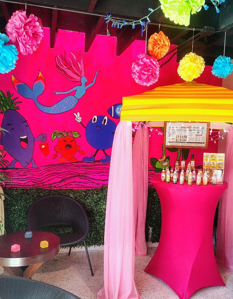 Colorful decorations with a pink background, hanging paper pom-poms, a mermaid illustration, and a pink and yellow booth with bottles.
