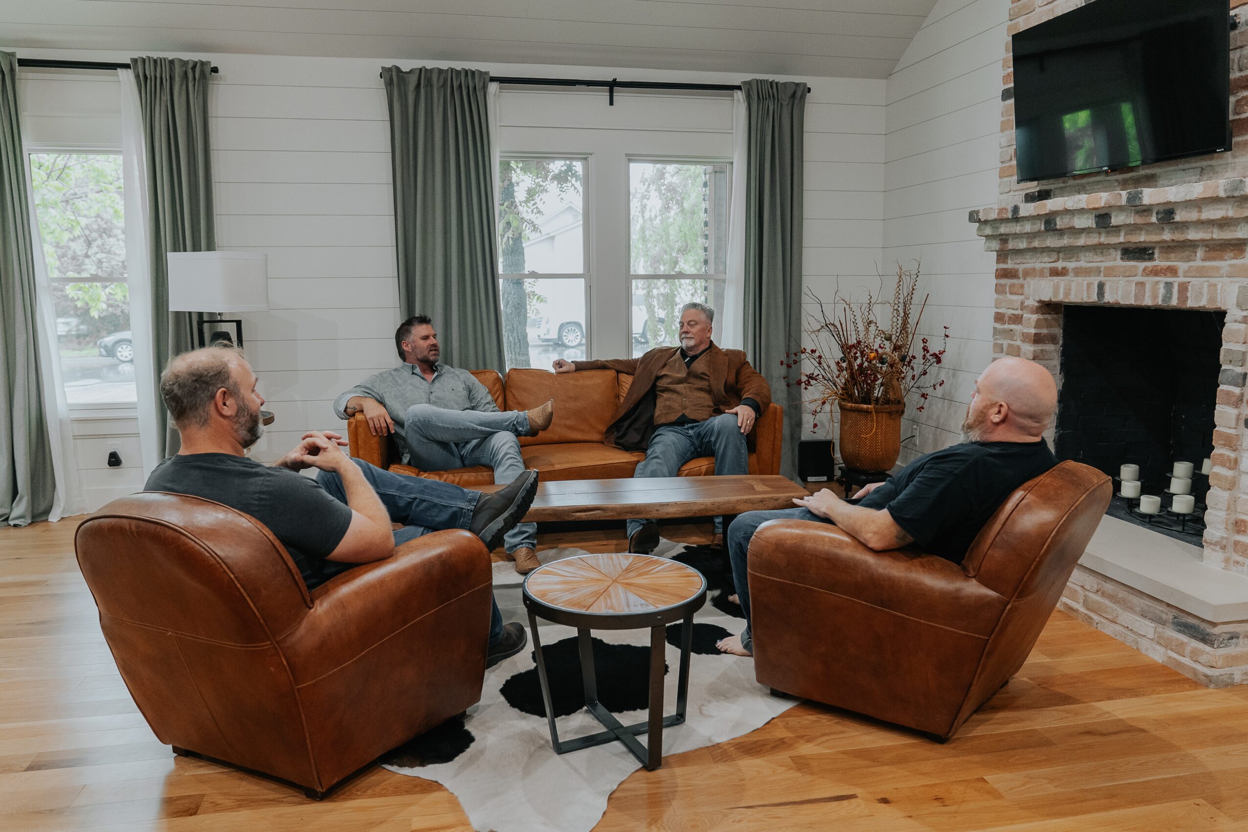 Four men sit in a living room with a fireplace, two on a sofa and two in leather chairs, talking and relaxing.