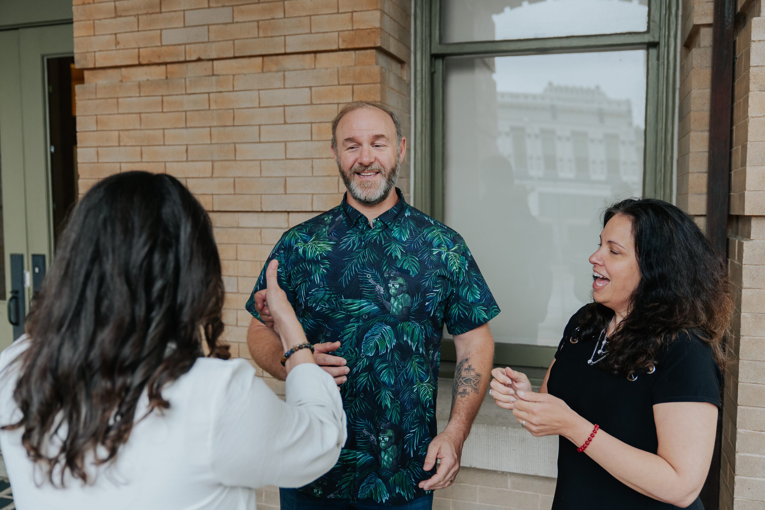 Three people smiling and talking outside a building with brick wall and window.