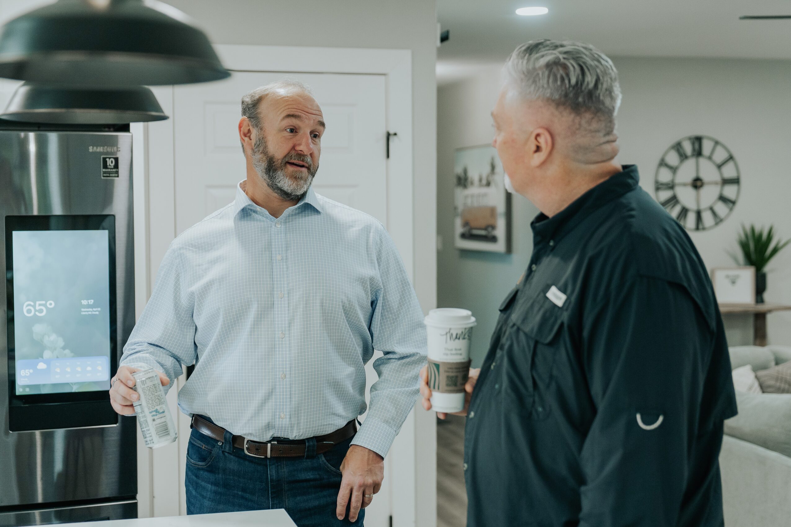Two men are talking in a modern kitchen, one holding a coffee cup, the other gesturing with his hand.