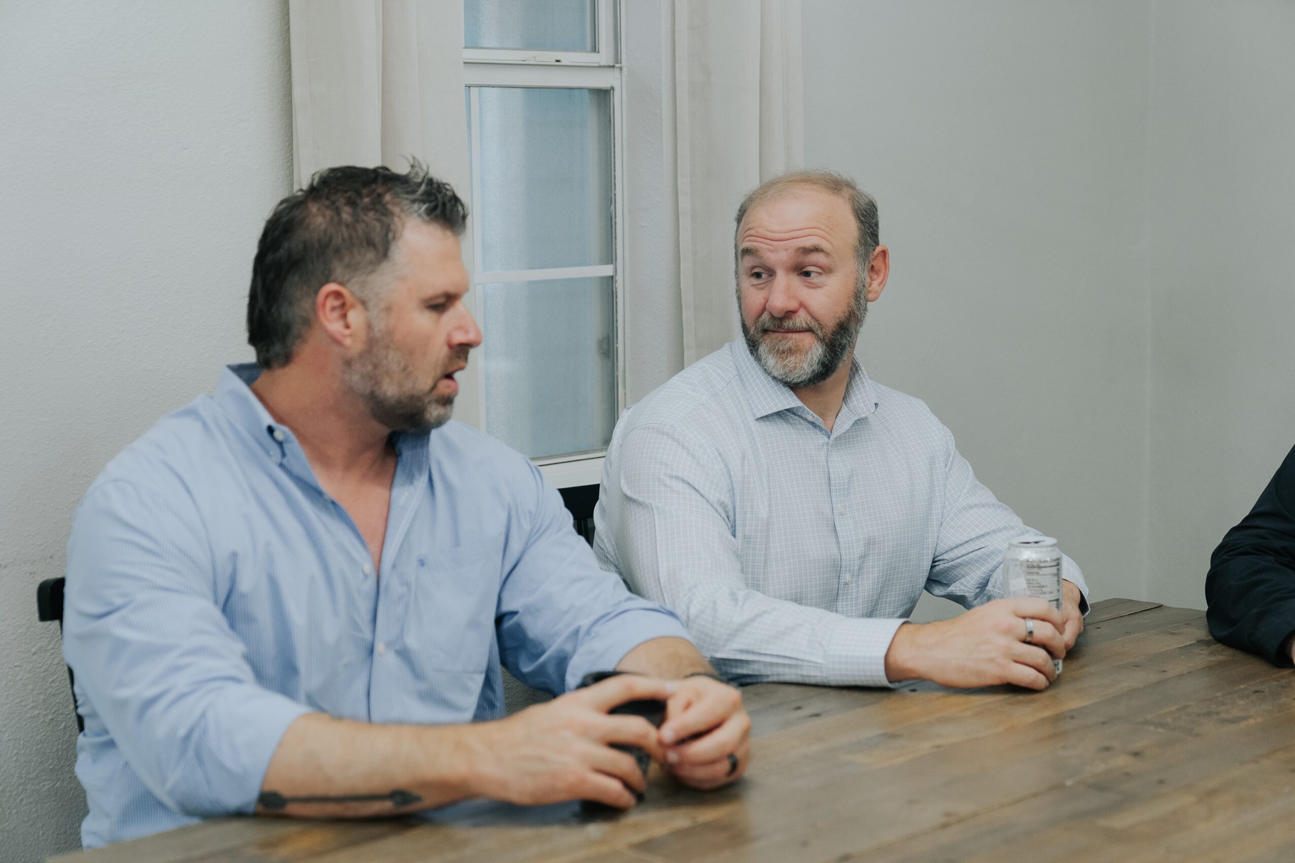 Two men sit at a wooden table, engaged in conversation, with a window behind them, in a light-colored room.