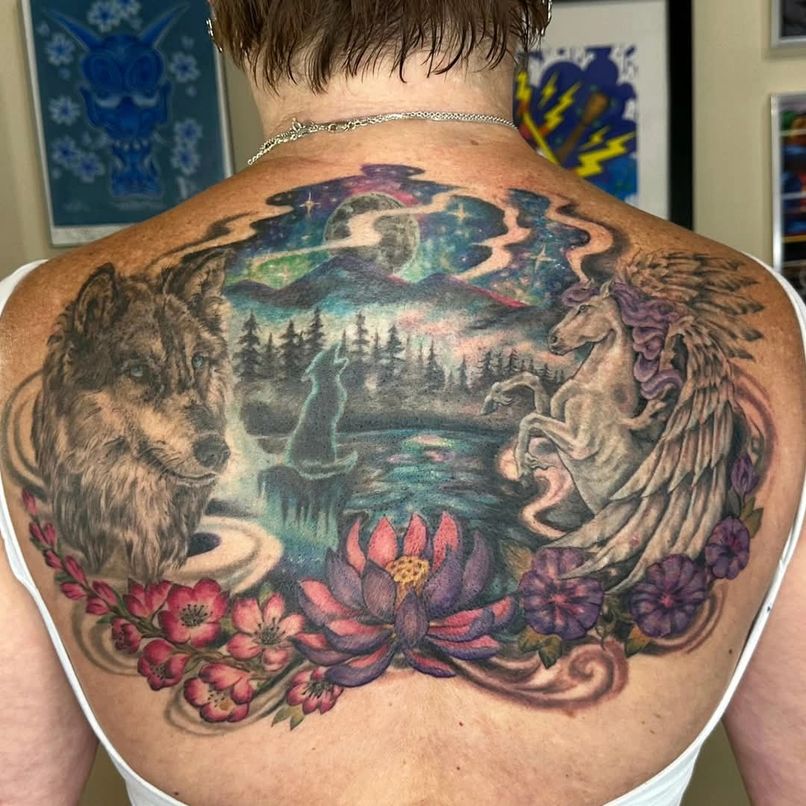 Colorful tattoo of a lion, wolf, unicorn, and flowers on a person's back, with a landscape background.
