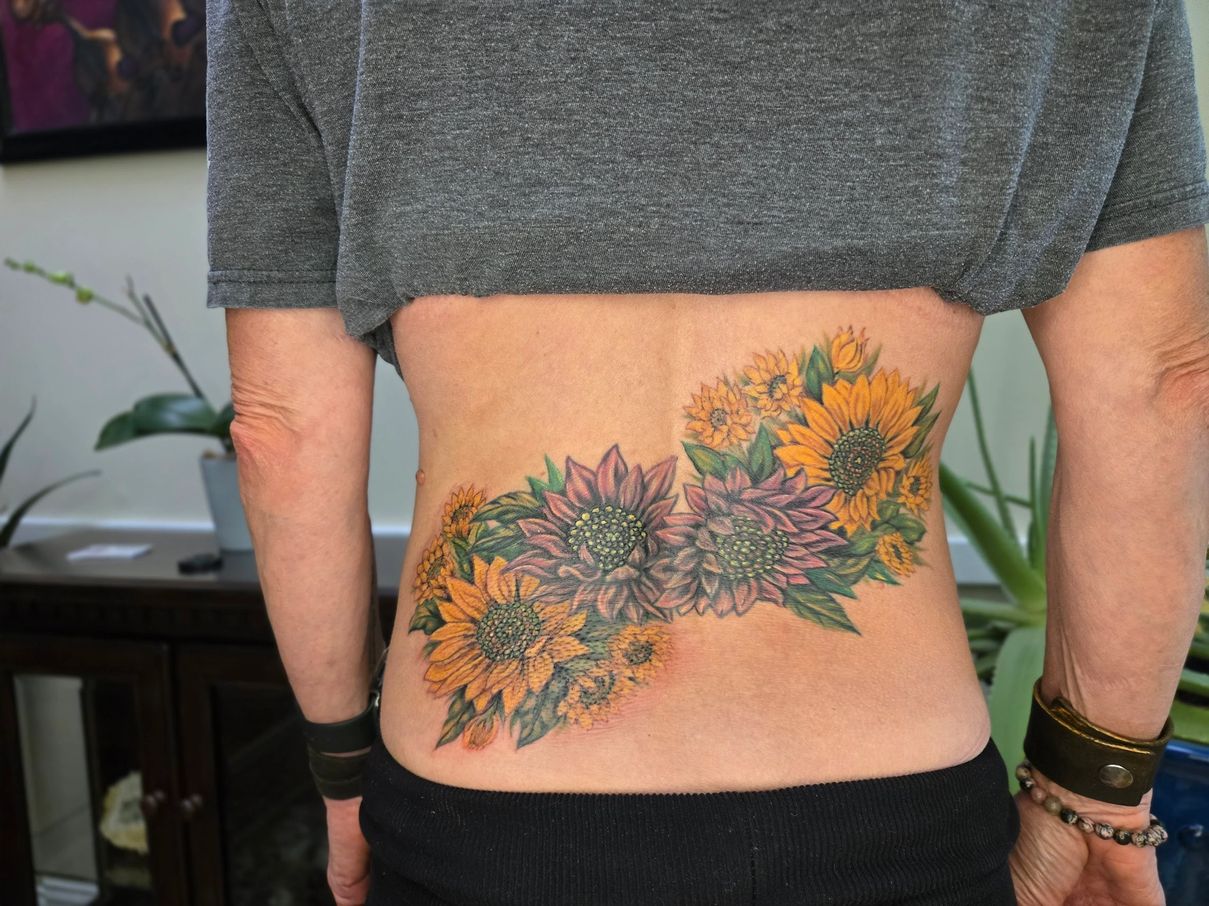 Person with a colorful floral tattoo on their side, wearing a gray shirt and black pants, standing indoors.