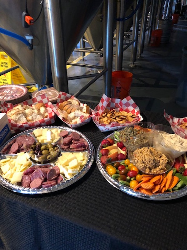 Assorted food items including cheese, meats, pickles, and fried chicken on a table with checkered paper baskets and vegetables.