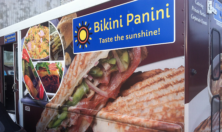 Food truck advertising a sandwich with lettuce, tomato, and grilled bread, and a sign reading 'Bikini Panini' with 'Taste the sunshine!'