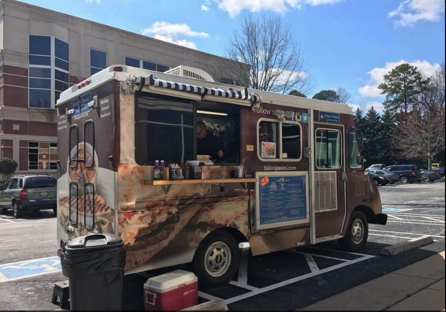 Food truck parked in a lot with a building and trees in the background, under a blue sky with clouds.