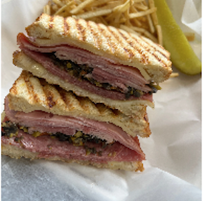 Stacked sandwich with ham, cheese, and olives, served with fries and a pickle spear in the background.