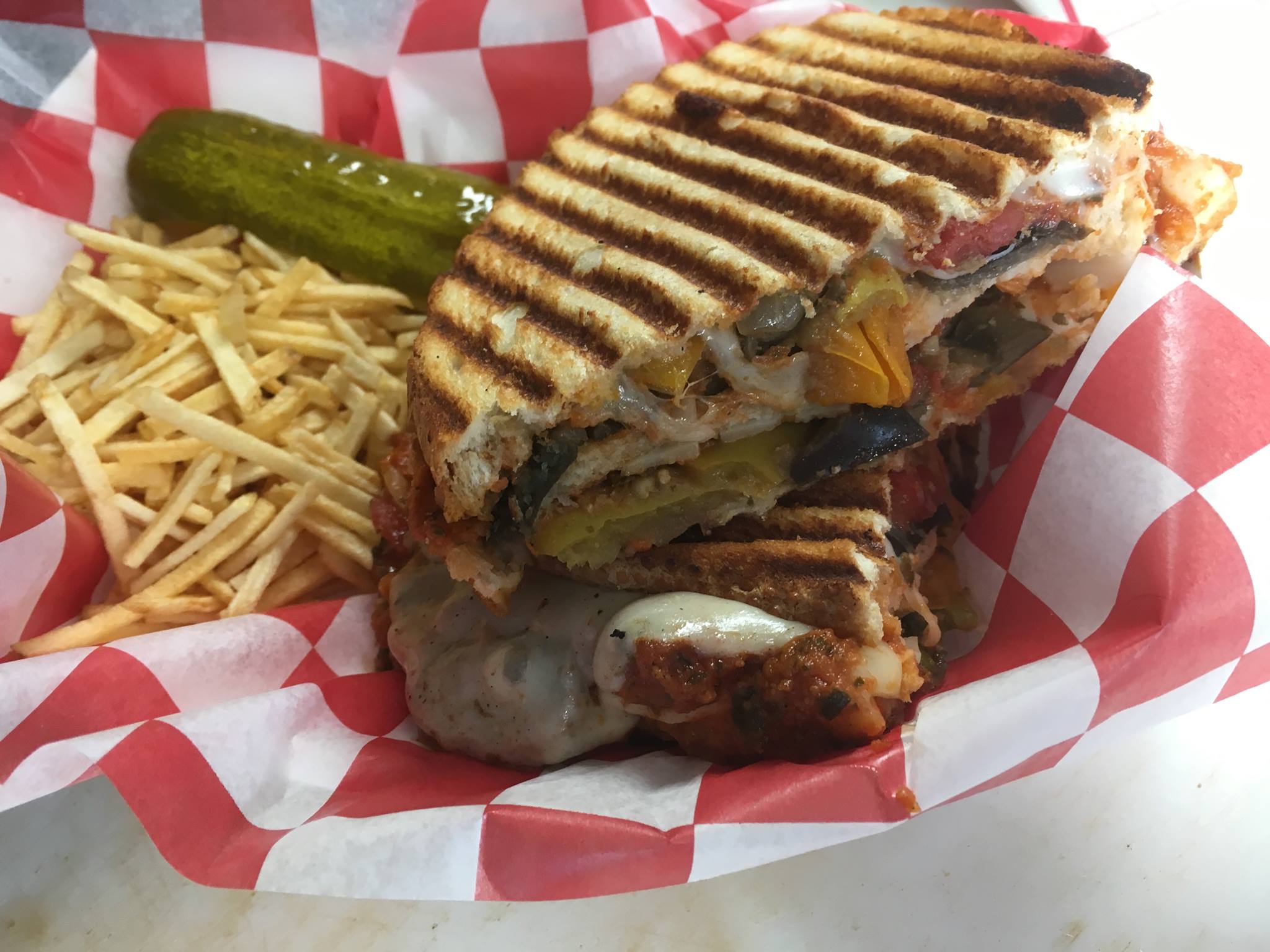 Grilled sandwich with meat, vegetables, and melted cheese, served with pickles, shredded cheese, and a side of fries on checkered paper.