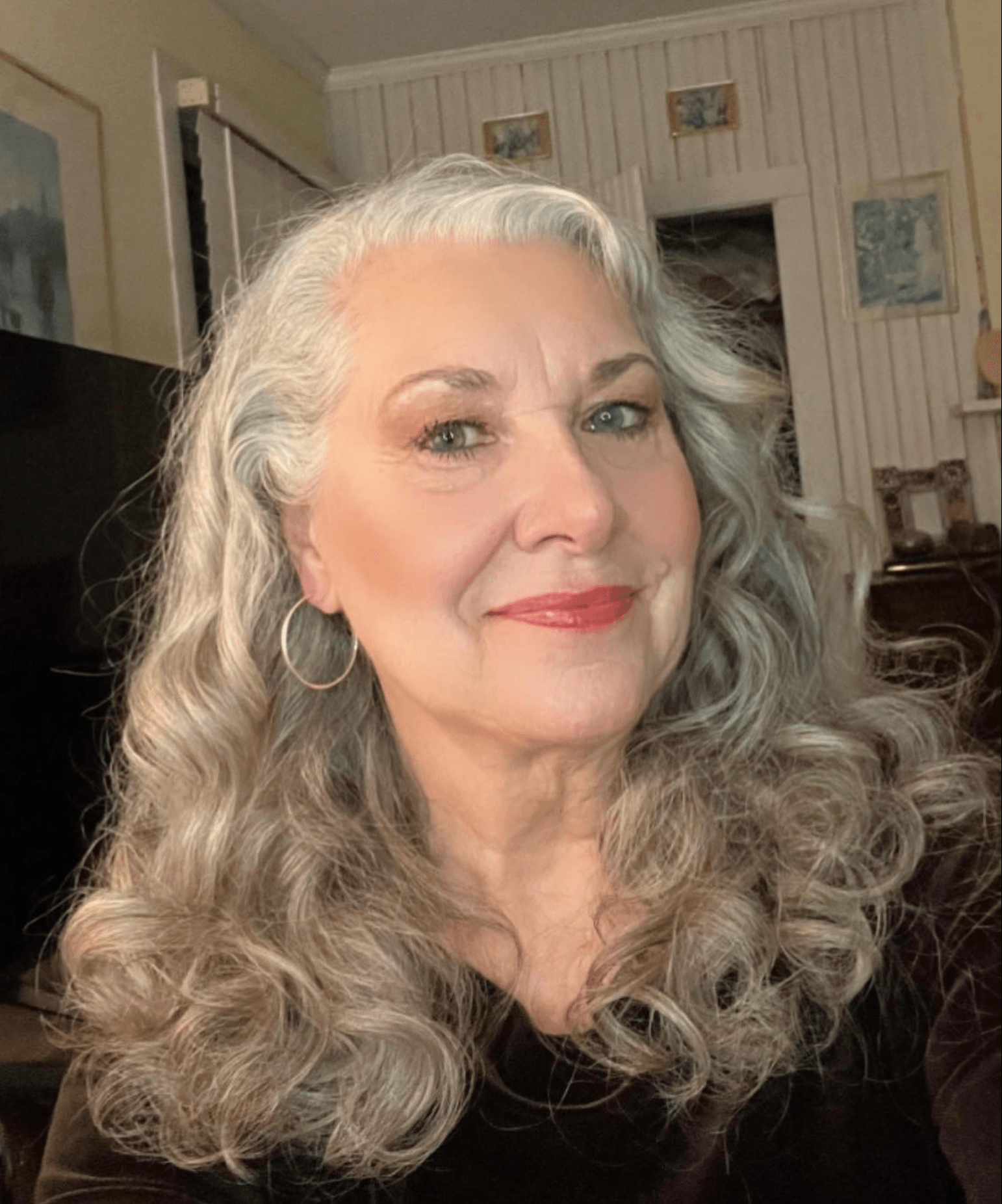 Portrait of a smiling woman with long, curly gray hair, wearing hoop earrings, in a cozy room with artwork on the wall.