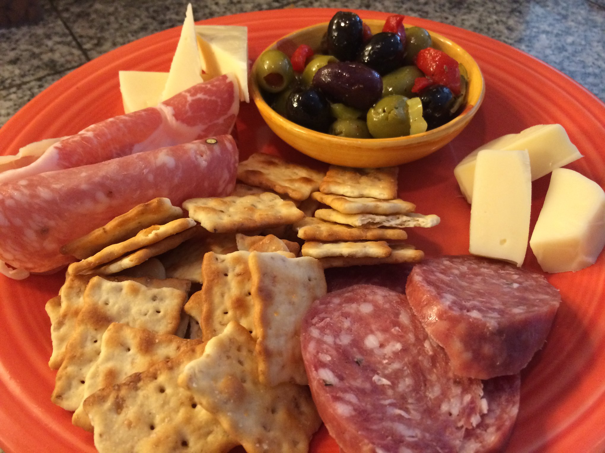 Assorted snacks on a red plate including cheese, crackers, cured meat, and a bowl of mixed olives.