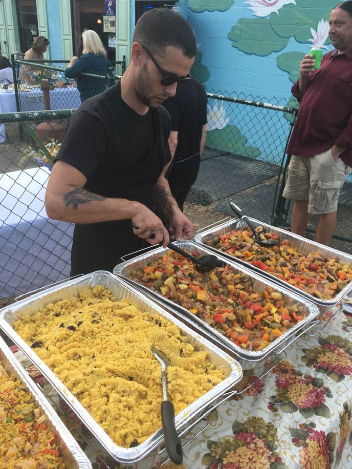 Man serving food from large trays at an outdoor event, with people in the background, a fence, and a colorful backdrop.