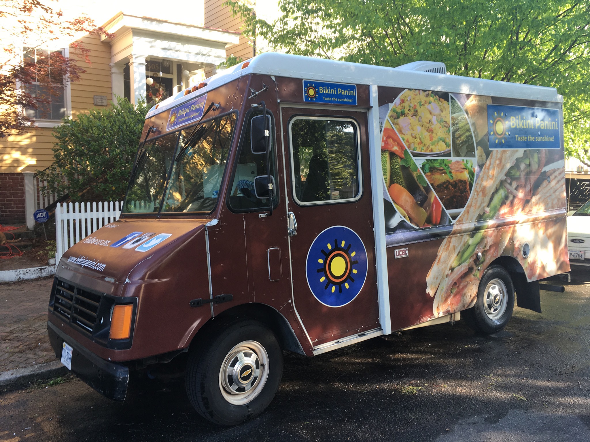 Food truck with colorful images of sandwiches and a sun logo parked on a street with trees and houses nearby.