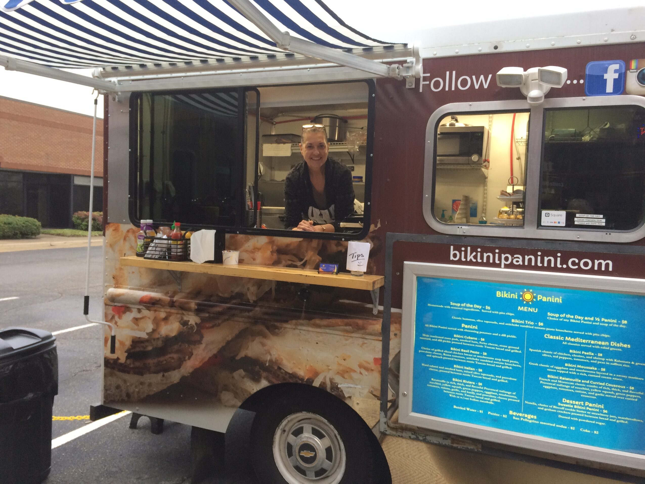 Person standing inside a food truck with a blue menu board outside, serving food outdoors.