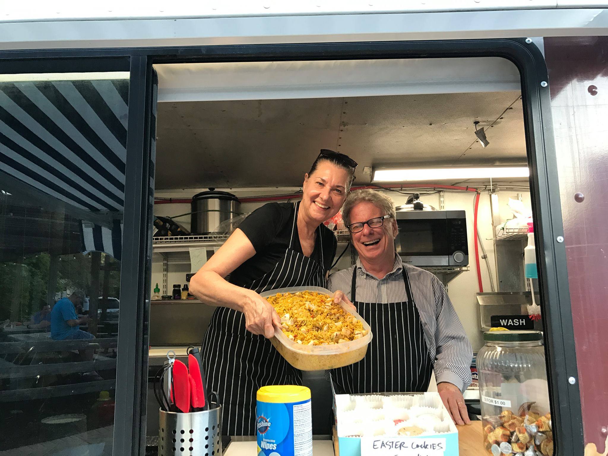 Two people in a food truck kitchen, smiling, holding a tray of food, with kitchen equipment in the background.