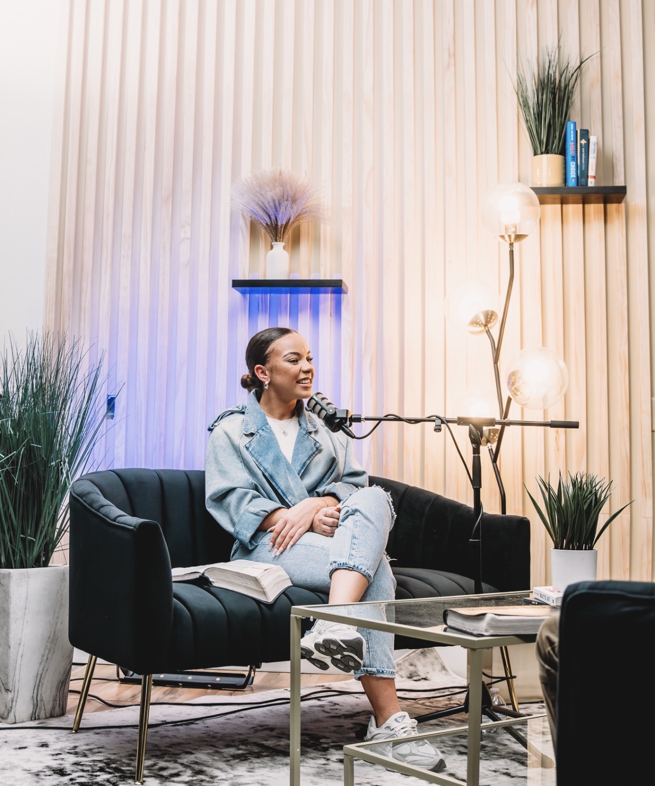 Woman sitting on a black sofa with a microphone, books, and plants in a modern room with vertical wooden panels.