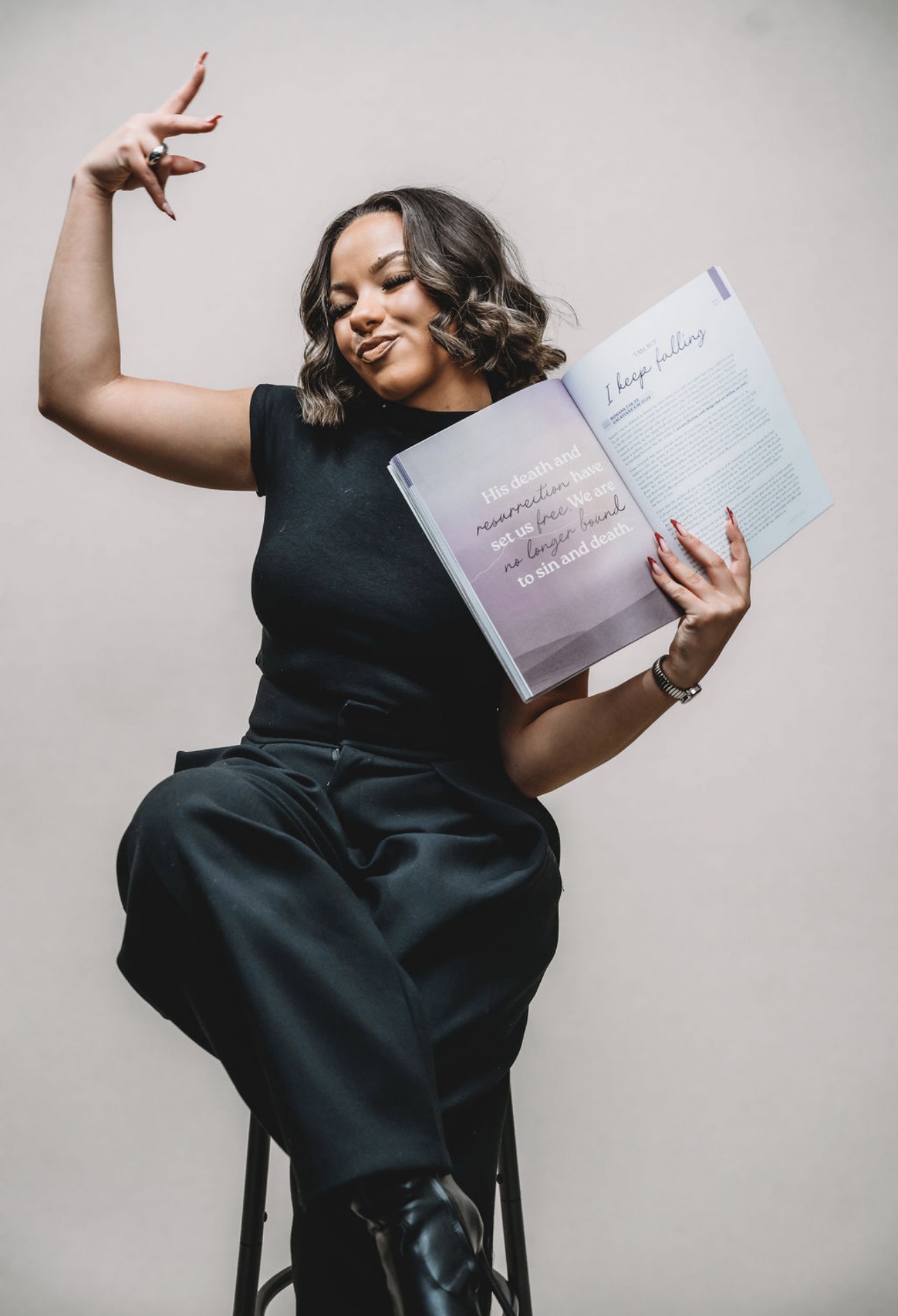 Woman sitting on a stool, reading a book, with one arm raised and a smile, against a plain background.