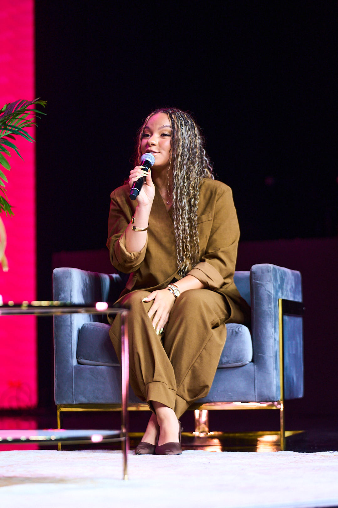 Woman with long curly hair sitting on a blue armchair, speaking into a microphone on stage.